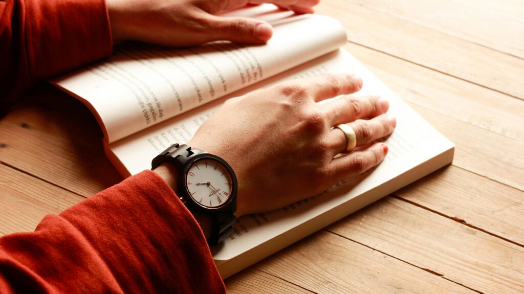 A person wearing a wristwatch resting hands on an open book on a wooden surface.